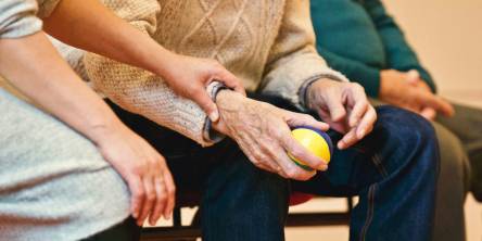 Three elderly people sitting on a bench. The man in the middle is holding a ball while the woman on the far right holds his forearm.