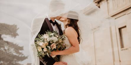 Bride and Groom kissing outdoors. The bride is holding a bouquet and their faces are covered by her veil. The groom's hand is on her hip facing the camera.